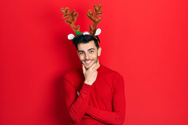 Şirin Noel geyiği boynuzları takan genç İspanyol adam kameraya kendinden emin bakıyor kollarını kavuşturmuş ve çenesini kaldırmış gülümsüyor. Olumlu düşünmek.  - Fotoğraf, Görsel