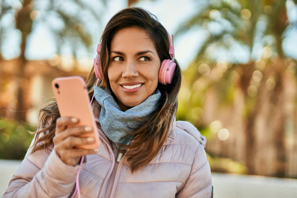 Joven mujer hispana sonriendo feliz usando teléfonos inteligentes y auriculares en la ciudad. - Foto, Imagen