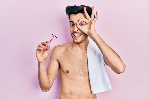 Молодий іспаномовний чоловік, що стоїть без сорочки, тримає бритву, усміхаючись, щасливий роблячи знак з рукою на око дивиться крізь пальці
  - Фото, зображення