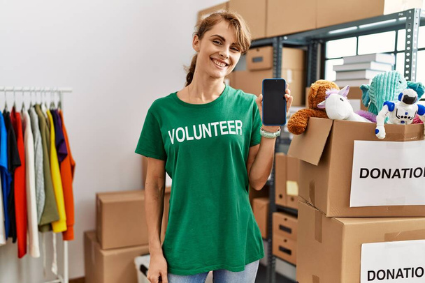 Jovem mulher voluntária caucasiana sorrindo feliz mostrando aplicativo de voluntariado no centro de caridade. - Foto, Imagem