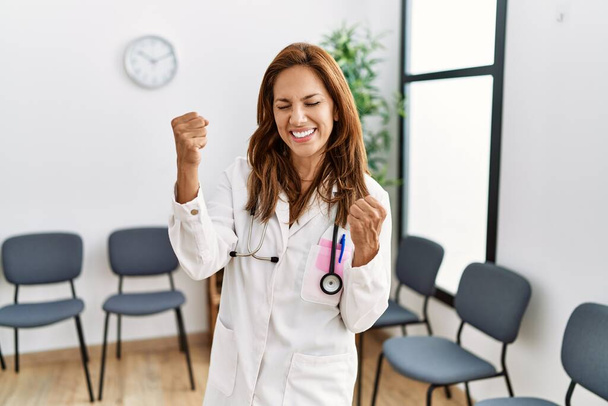 Középkorú spanyol orvos nő a váróteremben nagyon boldog és izgatott csinál győztes gesztus felemelt karokkal, mosolyog és sikoltozik a sikerért. ünnepség koncepciója.  - Fotó, kép