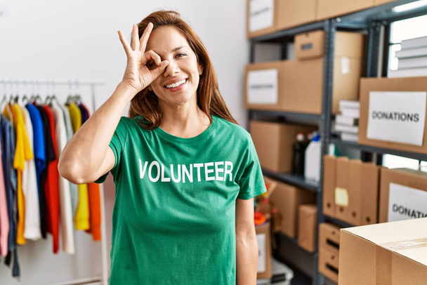 Eine hispanische Frau mittleren Alters trägt am Spendenstand ein freiwilliges T-Shirt und lächelt glücklich, als sie mit der Hand auf dem Auge durch die Finger schaut.  - Foto, Bild