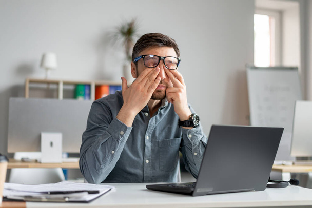 Volwassen mannelijke werknemer wrijven vermoeide geïrriteerde ogen, werken met laptop te veel, uitgeput van online baan op kantoor - Foto, afbeelding
