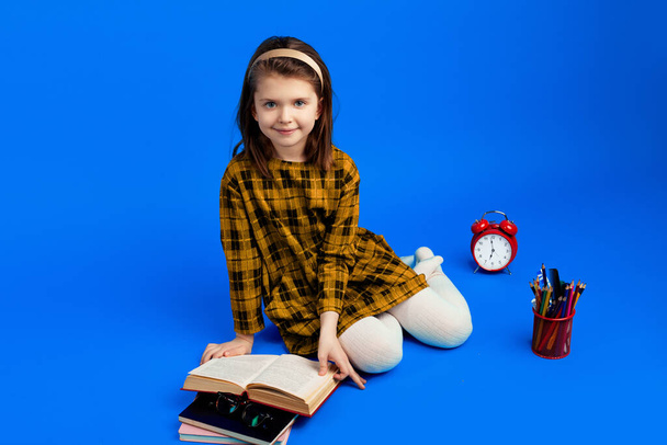 Onnellinen koulutyttö lukemassa kirjaa istuessaan sinistä taustaa vasten - Valokuva, kuva
