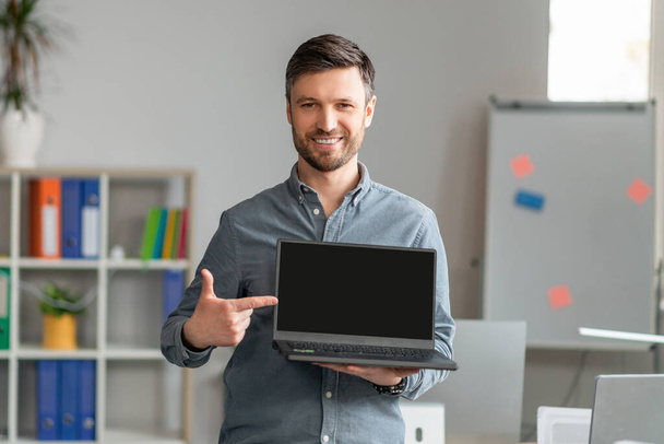 Portret szczęśliwego dojrzałego biznesmena wskazującego na laptop z czarnym ekranem, stojący w biurze, makieta - Zdjęcie, obraz