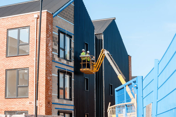 Bauarbeiter arbeiten am Teleskop-Boom-Lift und montieren isolierte Sandwich-Paneele an der Fassade eines neuen mehrstöckigen Wohnhauses. Sicher in der Höhe arbeiten - Foto, Bild