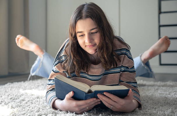 Söpö nuori nainen lukee kirjaa makatessaan lattialla huoneessaan kotona.. - Valokuva, kuva