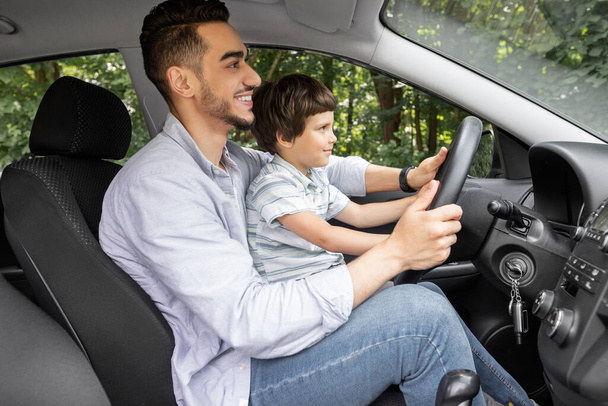 Happy millennial dad teaching little boy with steering wheel to drive car in summer, outdoor, profile - Photo, image