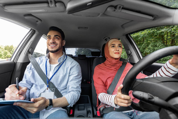 Χαμογελώντας χιλιετή αραβική γυναίκα σε hijab λαμβάνοντας δοκιμή οδήγησης με τον μέντορα άνδρα, κυρία στο τιμόνι οδήγηση στο αυτοκίνητο - Φωτογραφία, εικόνα
