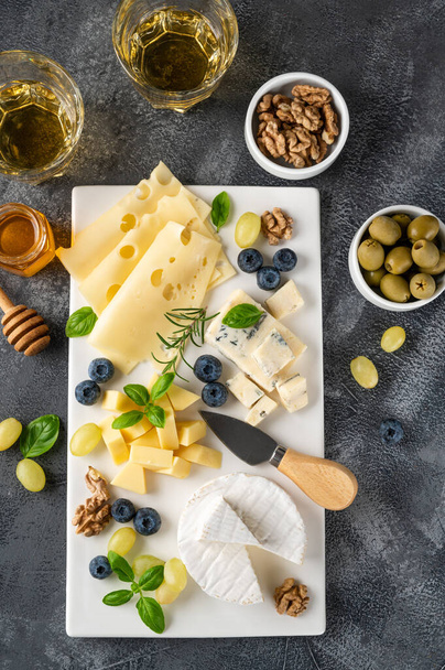 Assiette au fromage avec une variété de fromages, miel, raisins, noix, olives, bleuets et herbes fraîches sur un fond de béton. Une collation festive - Photo, image