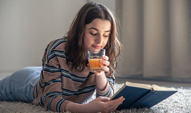 Молода жінка читає книгу і п'є апельсиновий сік, лежачи на підлозі в кімнаті
. - Фото, зображення