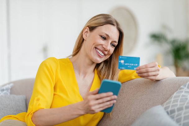 Joven rubia europea alegre en ropa amarilla con teléfono inteligente y tarjetas de crédito de compras en línea - Foto, imagen