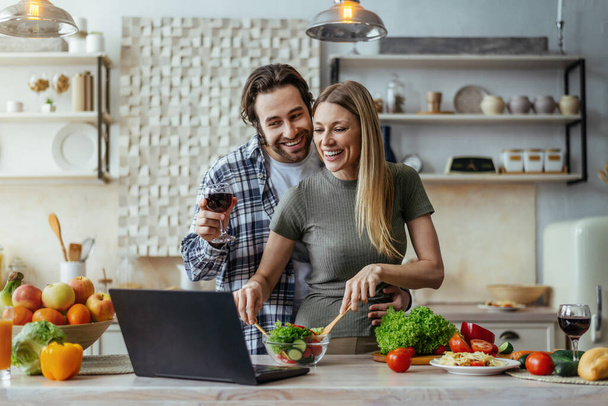 Zufriedener kaukasischer Millennial-Ehemann mit Stoppeln und Frau, die zusammen kochen, Wein trinken und am Laptop reden - Foto, Bild
