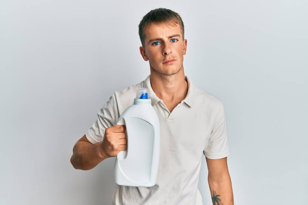 Jovem caucasiano segurando detergente garrafa pensando atitude e expressão sóbria olhando auto confiante  - Foto, Imagem