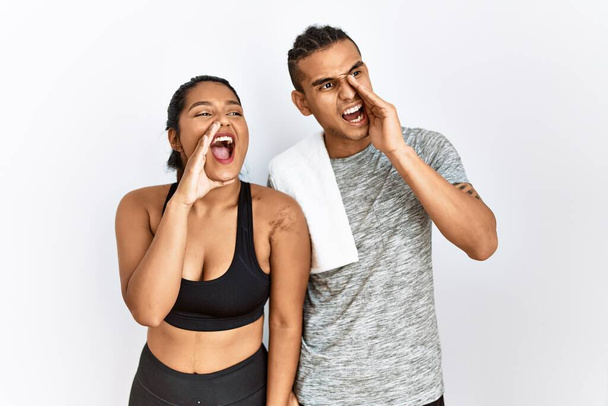 Молода латинська пара носить спортивний одяг, що стоїть над ізольованим фоном, кричить і кричить голосно на бік з рукою на рот. концепція спілкування
.  - Фото, зображення