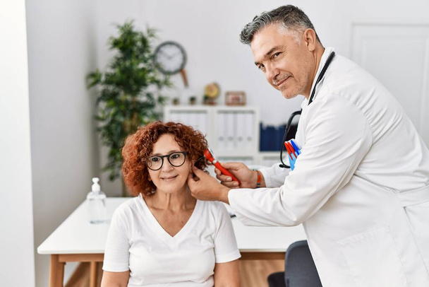 Middelbare leeftijd man en vrouw in doktersuniform onderzoeken oor met medisch consult in kliniek - Foto, afbeelding