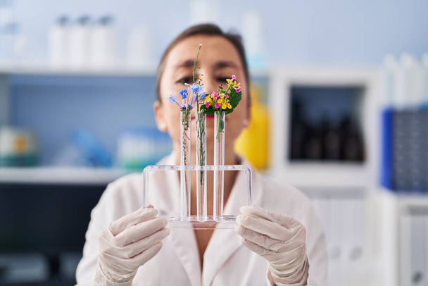 Middle age woman scientist holding test tubes with flower at laboratory - Фото, изображение