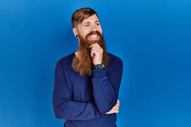 Рудий чоловік з довгою бородою носить випадковий синій светр на синьому фоні з рукою на підборідді, думаючи про питання, стомлюючий вираз. усміхнене і вдумливе обличчя. концепція сумніву
.  - Фото, зображення