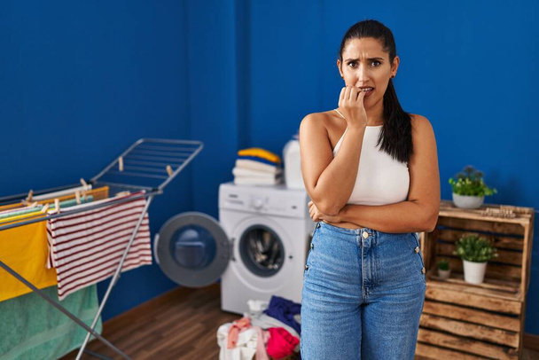 Young hispanic woman at laundry room looking stressed and nervous with hands on mouth biting nails. anxiety problem.  - Photo, Image