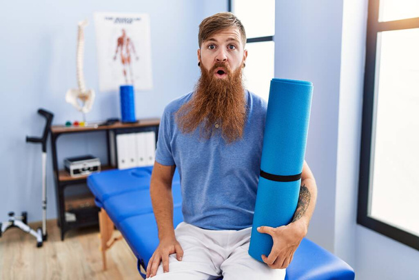 Redhead man with long beard holding yoga mat at rehabilitation clinic scared and amazed with open mouth for surprise, disbelief face  - Photo, image