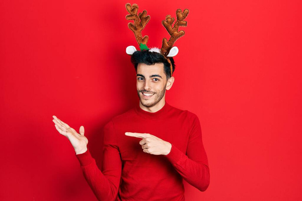 jovem hispânico homem vestindo bonito Natal renas chifres espantado e sorrindo para a câmera ao apresentar com a mão e apontando com o dedo.  - Foto, Imagem