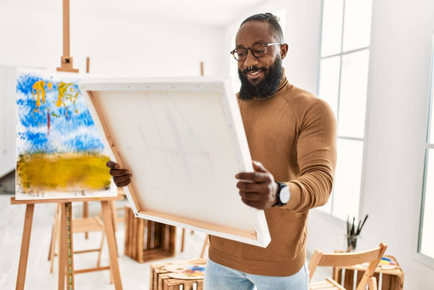 młody afrykański artysta amerykański człowiek uśmiecha szczęśliwy trzymając płótno w studio sztuki. - Zdjęcie, obraz