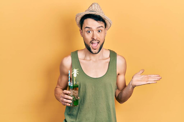 Joven hispano con sombrero de verano bebiendo mojito celebrando el logro con sonrisa feliz y expresión ganadora con la mano levantada  - Foto, imagen