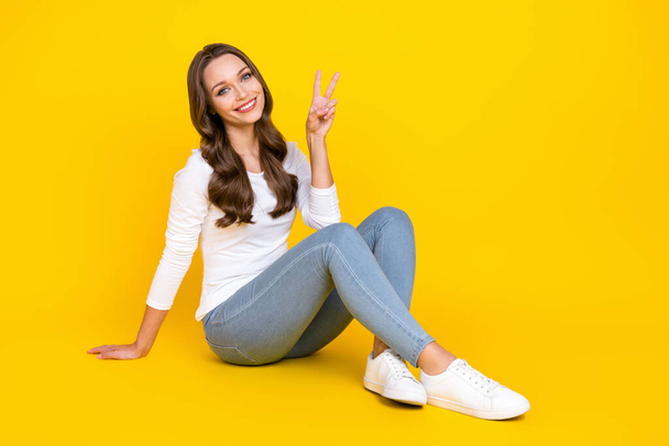 Photo of confident lovely friendly lady sit floor show v-sign wear white shirt isolated yellow color background - Foto, Imagem