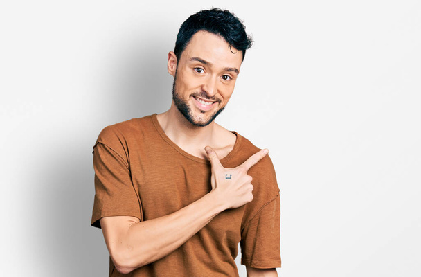 Homme hispanique à la barbe portant un t-shirt décontracté souriant pointant joyeusement avec la main et le doigt sur le côté  - Photo, image
