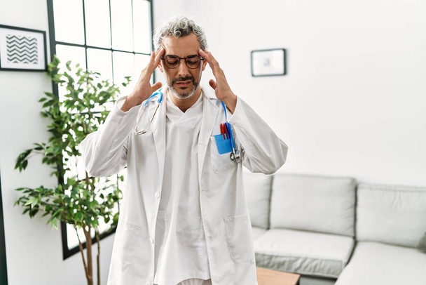 Hombre hispano de mediana edad vistiendo uniforme médico y estetoscopio en la sala de espera con la mano en la cabeza para el dolor en la cabeza porque el estrés. padecer migraña.  - Foto, Imagen