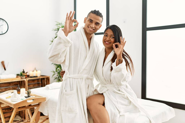 Young latin couple wearing towel standing at beauty center smiling positive doing ok sign with hand and fingers. successful expression.  - Foto, afbeelding