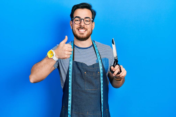 Handsome caucasian man with beard dressmaker designer wearing atelier apron holding scissors smiling happy and positive, thumb up doing excellent and approval sign  - Foto, imagen