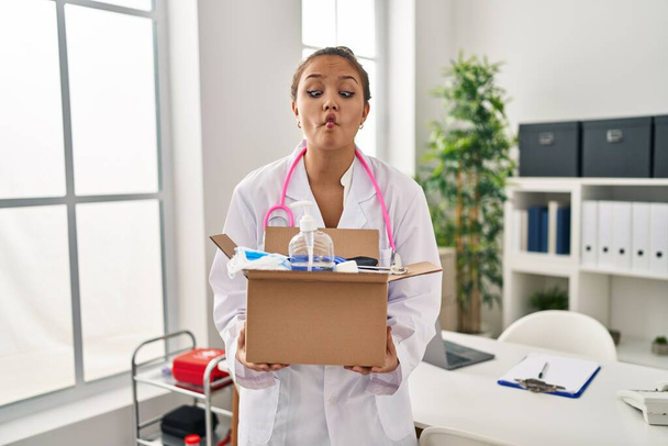 Young hispanic doctor holding box with medical items making fish face with mouth and squinting eyes, crazy and comical.  - Фото, изображение