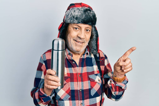 Handsome mature handyman wearing winter hat with ear flaps holding thermo smiling happy pointing with hand and finger to the side  - Photo, Image