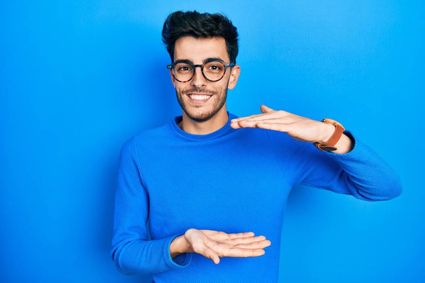 Young hispanic man wearing casual clothes and glasses gesturing with hands showing big and large size sign, measure symbol. smiling looking at the camera. measuring concept.  - Zdjęcie, obraz