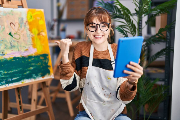 Young beautiful artist woman doing video call with tablet pointing thumb up to the side smiling happy with open mouth  - Photo, Image