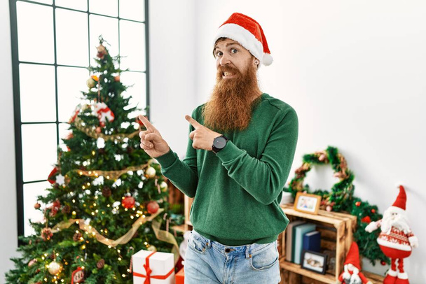 Redhead man with long beard wearing christmas hat by christmas tree pointing aside worried and nervous with both hands, concerned and surprised expression  - Foto, afbeelding