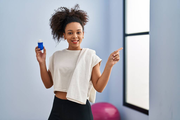 Giovane donna afroamericana con indosso abbigliamento sportivo e asciugamano che tiene deodorante sorridente felice indicando con mano e dito di lato  - Foto, immagini