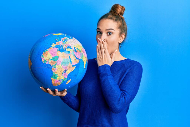 Mujer rubia joven sosteniendo bola mundial cubriendo la boca con la mano, conmocionada y temerosa de equivocarse. expresión sorprendida  - Foto, Imagen