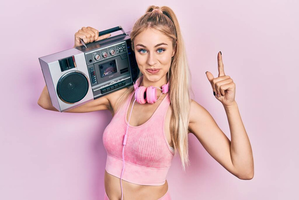 Young beautiful caucasian woman wearing workout clothes and holding boombox smiling with an idea or question pointing finger with happy face, number one  - Fotoğraf, Görsel