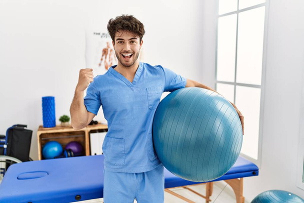 Hispanic physiotherapy man working at pain recovery clinic screaming proud, celebrating victory and success very excited with raised arms  - Fotografie, Obrázek