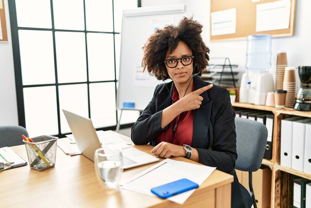 Femme afro-américaine avec des cheveux afro travaillant au bureau portant casque opérateur pointant avec le doigt de la main sur le côté montrant la publicité, visage sérieux et calme  - Photo, image