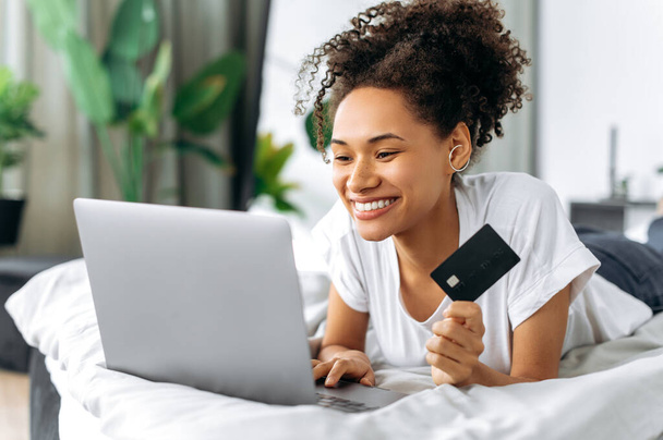 Online winkelen, e-commerce. Positieve mooie jonge Afrikaanse Amerikaanse vrouw liggend op een bed, gebruikt laptop en creditcard om te betalen voor online aankopen en levering, voert gegevens, kijkt naar het scherm, glimlacht - Foto, afbeelding