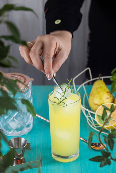 Real woman preparing Pear Collins Cocktail in highball glass or mocktails surrounded by ingredients and bar tools on turquoise table surface. Ready for drinking. - Фото, изображение