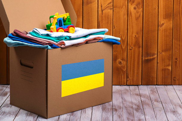 Κιβώτιο δωρεών με ανθρωπιστική βοήθεια με παιδικά πράγματα και παιχνίδια για Ουκρανούς πρόσφυγες. Φιλανθρωπία και βοήθεια σε φτωχούς και άπορους ανθρώπους. Μείνε με την Ουκρανία. Ουκρανική σημαία στο κουτί με δωρεά. - Φωτογραφία, εικόνα