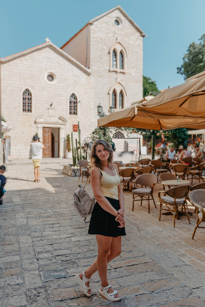 Girl Tourist Walking Through Ancient Narrow Street On A Beautiful Summer Day In MEDITERRANEAN MEDIEVAL CITY, OLD TOWN BUDVA, MONTENEGRO. Young Beautiful Cheerful Woman Walking On Old Street At - Foto, Imagen