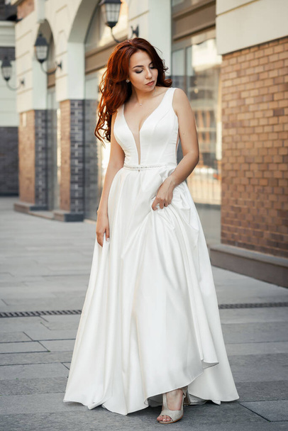 Refined feminine lady with red hair in a white dress. A happy and satisfied bride poses on a city street. Summer mood - Valokuva, kuva