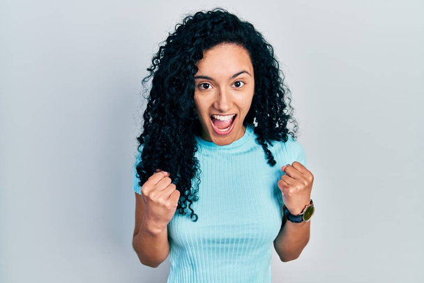 Junge hispanische Frau mit lockigem Haar in lässigem blauem T-Shirt, die überrascht und erstaunt den Erfolg mit erhobenen Armen und offenen Augen feiert. Siegerkonzept.  - Foto, Bild