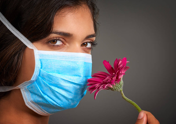 I cant seem to smell anything. Closeup of a young woman with a doctors mask on looking at the camera and smelling a flower. - Photo, Image