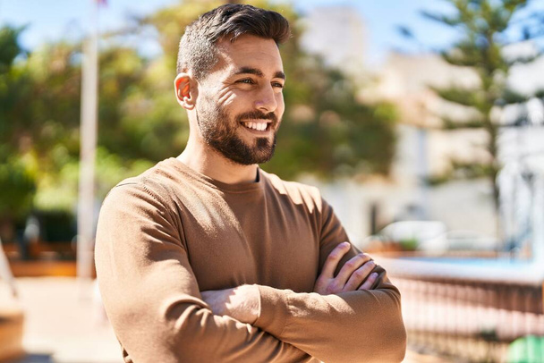 Fiatal spanyol férfi mosolyogva magabiztosan áll keresztbe tett kézzel a parkban. - Fotó, kép